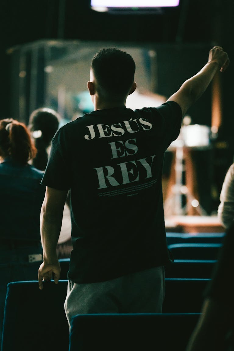 A man in a church raises his hand in worship wearing a shirt that says 'Jesus es Rey'.